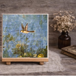 May include: A square decorative tile featuring a printed image of a small brown bird in flight against a textured blue background with green foliage and yellow flowers, supported by a light-colored wooden stand with a rectangular base.