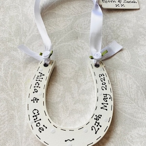 May include: White horseshoe with black lettering, reading "Alicia & Chloe" on one side and "27th May 2023" on the other. It is decorated with white satin ribbon and small white flowers. The horseshoe is accompanied by a white tag with black lettering that reads "Love from Nicky, Andrew Björn & Sarah Xx".