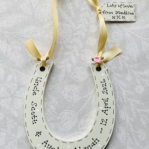 May include: A cream-colored wooden horseshoe decoration featuring white polka dots and black text reading Uncle Scott & Auntie Alanah ~ 12 April 2026. It includes a yellow ribbon, two small fabric roses, and a tag saying Lots of love from Madison xxx.