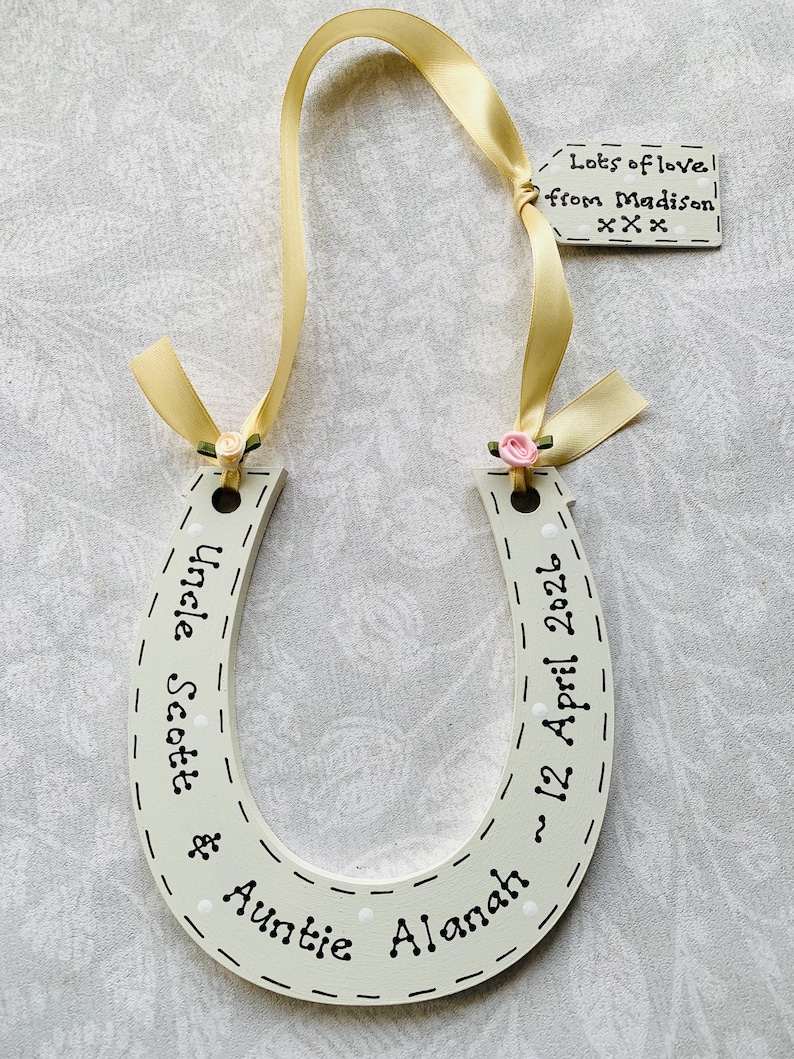 May include: A cream-colored wooden horseshoe decoration featuring white polka dots and black text reading Uncle Scott & Auntie Alanah ~ 12 April 2026. It includes a yellow ribbon, two small fabric roses, and a tag saying Lots of love from Madison xxx.