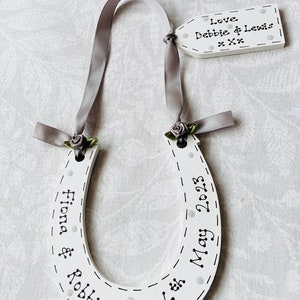 May include: A white wooden horseshoe ornament featuring black text that reads Fiona & Robbie 6th May 2023, decorated with grey ribbon, small grey fabric roses, and a matching rectangular tag inscribed with Love Debbie & Lewis xxx.