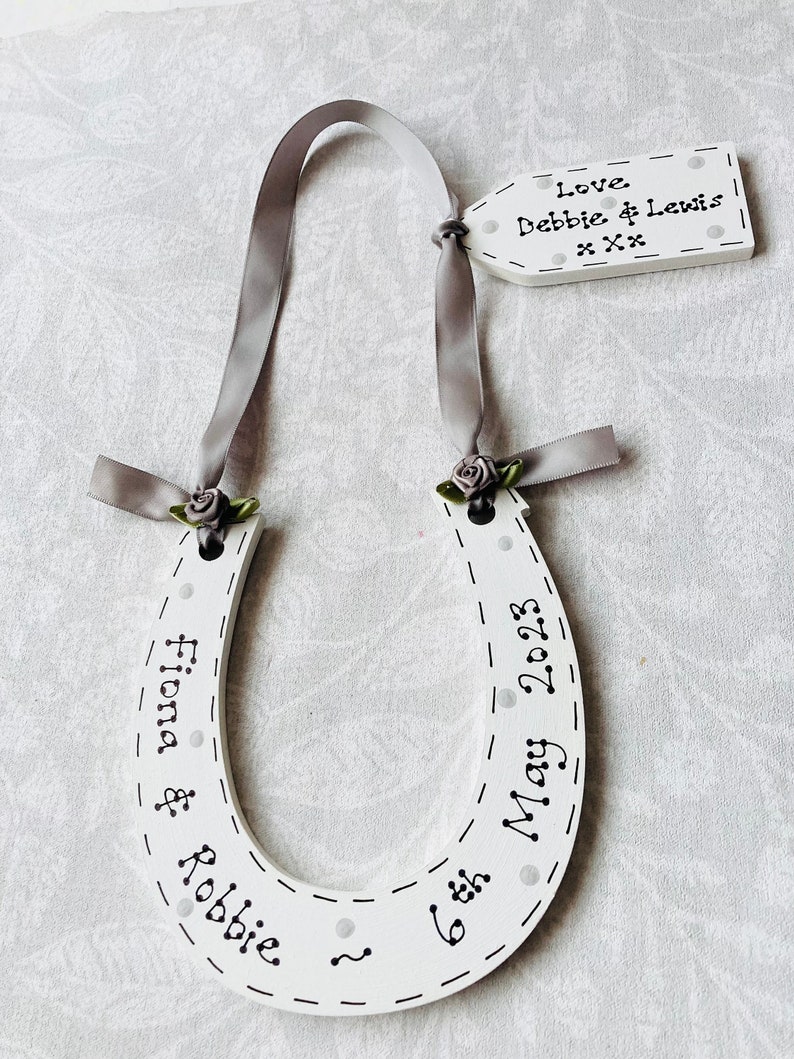 May include: A white wooden horseshoe ornament featuring black text that reads Fiona & Robbie 6th May 2023, decorated with grey ribbon, small grey fabric roses, and a matching rectangular tag inscribed with Love Debbie & Lewis xxx.
