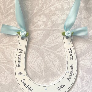 May include: A white horseshoe-shaped ornament featuring the text Mummy & Daddy and 7th August 2026. It is decorated with light blue satin ribbons and small light blue fabric roses with green leaves attached to the top ends of the horseshoe.