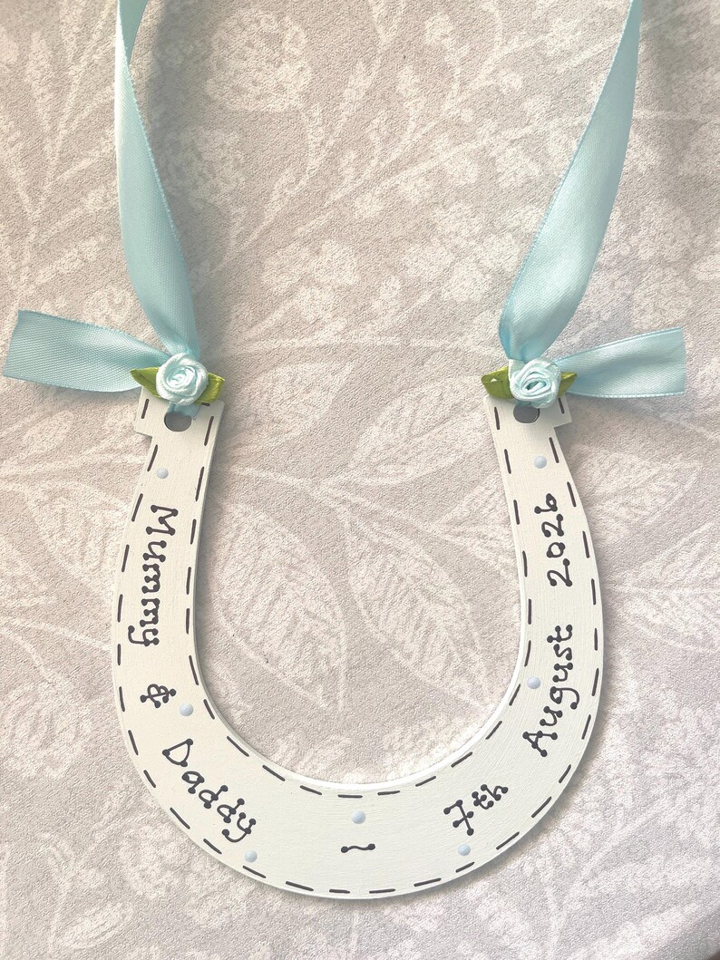 May include: A white horseshoe-shaped ornament featuring the text Mummy & Daddy and 7th August 2026. It is decorated with light blue satin ribbons and small light blue fabric roses with green leaves attached to the top ends of the horseshoe.