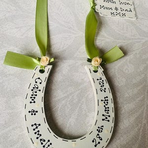 May include: A white wooden horseshoe ornament featuring black text reading Calum & Amelia and 28th March 2026. It is decorated with light green satin ribbons, small cream-colored fabric roses, and a small white tag reading With love, Mum & Dad xxx.