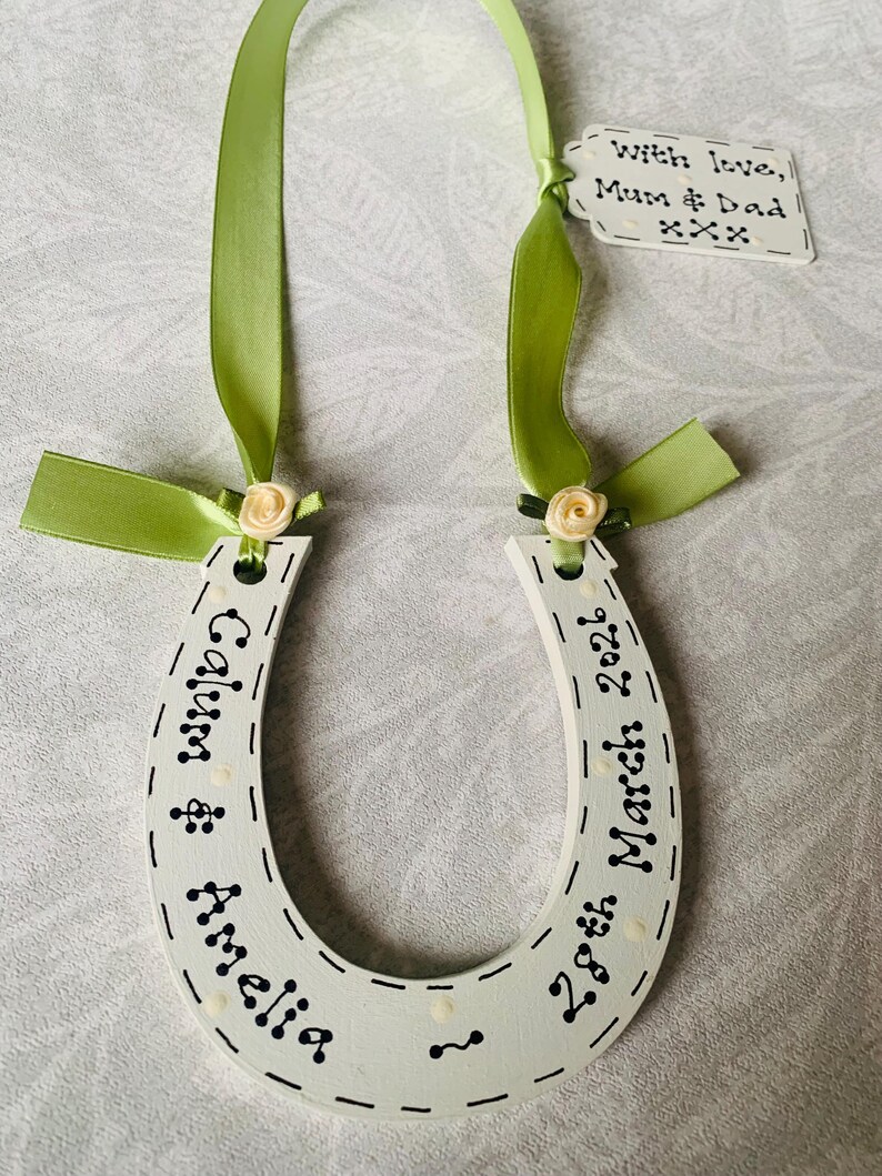 May include: A white wooden horseshoe ornament featuring black text reading Calum & Amelia and 28th March 2026. It is decorated with light green satin ribbons, small cream-colored fabric roses, and a small white tag reading With love, Mum & Dad xxx.