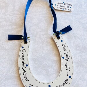 May include: A white wooden horseshoe decoration featuring dark blue ribbon and hand-painted text that reads Congratulations, 15th April 2023, and Stuart & Bronwyn. A small matching tag attached to the ribbon reads Lots of love Sidney & Spencer xx.
