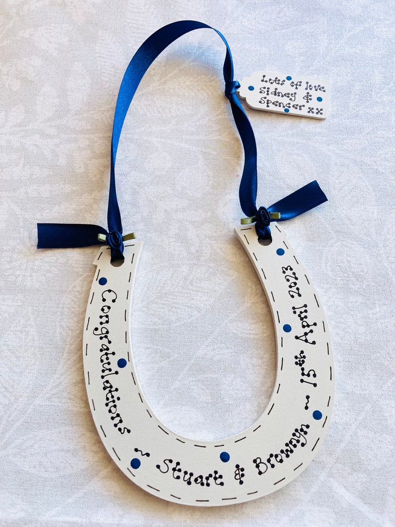 May include: A white wooden horseshoe decoration featuring dark blue ribbon and hand-painted text that reads Congratulations, 15th April 2023, and Stuart & Bronwyn. A small matching tag attached to the ribbon reads Lots of love Sidney & Spencer xx.