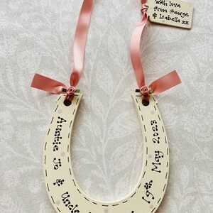 May include: A cream-colored wooden horseshoe decoration featuring black hand-lettered text reading "Auntie Jo & Uncle Keith" and "19th May 2023". It is adorned with a pink satin ribbon, small fabric roses, and a small rectangular wooden tag reading "with love from George & Isabella xx".