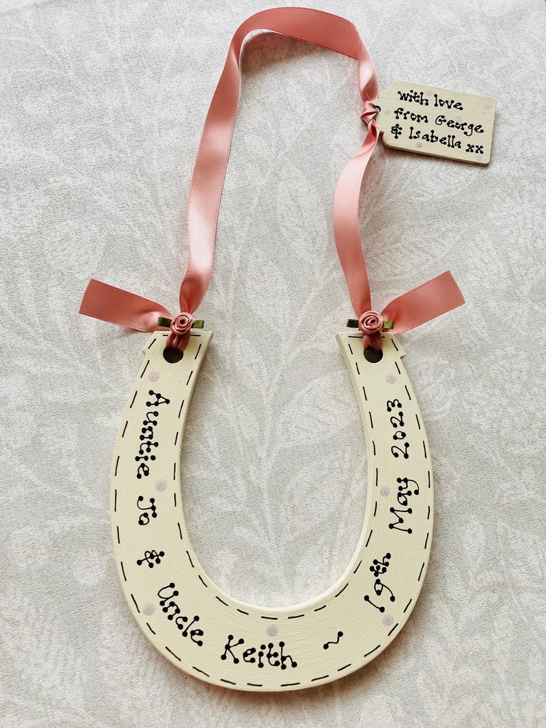 May include: A cream-colored wooden horseshoe decoration featuring black hand-lettered text reading "Auntie Jo & Uncle Keith" and "19th May 2023". It is adorned with a pink satin ribbon, small fabric roses, and a small rectangular wooden tag reading "with love from George & Isabella xx".