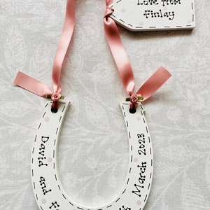 May include: A white wooden horseshoe ornament decorated with light pink polka dots and black dashed lines. It features the text "David and Faye" and "11th March 2023" with a pink ribbon and a small tag reading "Love from Finlay" attached.