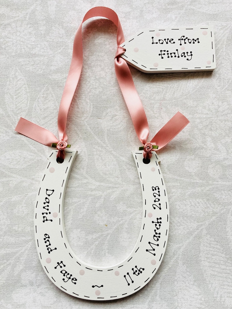 May include: A white wooden horseshoe ornament decorated with light pink polka dots and black dashed lines. It features the text "David and Faye" and "11th March 2023" with a pink ribbon and a small tag reading "Love from Finlay" attached.