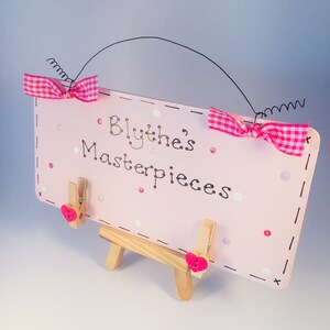Könnte beinhalten: Ein rosa Holzschild mit dem Text "Blythe's Masterpieces" und zwei rosa Schleifen. Das Schild steht auf einem kleinen Holzständer und hat zwei Wäscheklammern mit rosa Herzen.