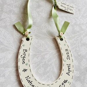 May include: A cream-colored wooden horseshoe decoration featuring black text that reads Bryce & Natasha and 29th April 2023. It is adorned with light green ribbon, small cream fabric roses, and a small tag reading Lots of love from Maya xx.