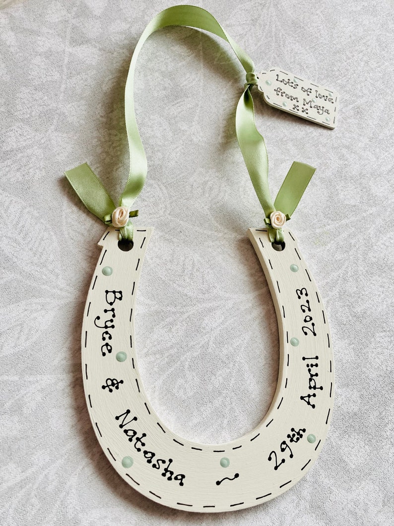 May include: A cream-colored wooden horseshoe decoration featuring black text that reads Bryce & Natasha and 29th April 2023. It is adorned with light green ribbon, small cream fabric roses, and a small tag reading Lots of love from Maya xx.