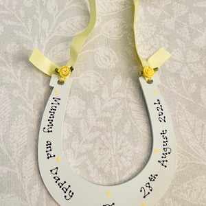 May include: A white horseshoe-shaped wooden ornament featuring yellow ribbon accents, small yellow fabric roses, and black text reading Mummy and Daddy 28th August 2026. A small attached tag displays the text Lots of love Clara & Tomas x.