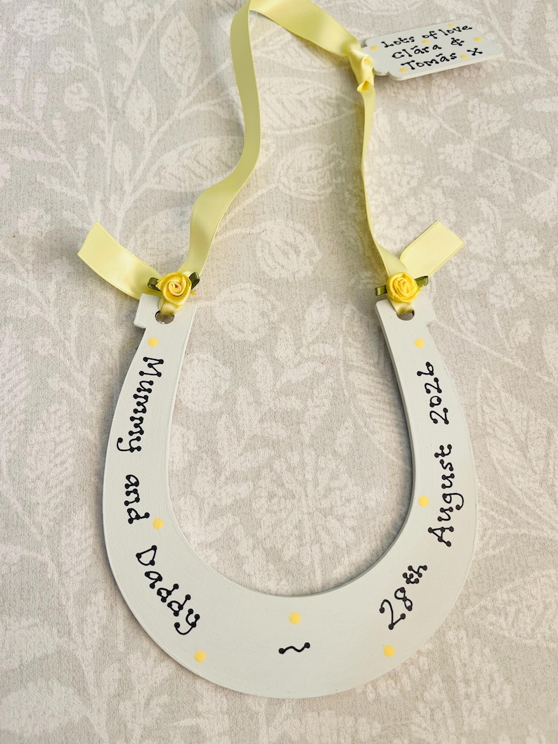 May include: A white horseshoe-shaped wooden ornament featuring yellow ribbon accents, small yellow fabric roses, and black text reading Mummy and Daddy 28th August 2026. A small attached tag displays the text Lots of love Clara & Tomas x.