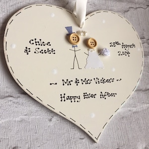 May include: White heart-shaped wooden sign with a hand-drawn illustration of a bride and groom, the text "Chloe & Scott", "25th August 2014", "Mr & Mrs Vickers", and "Happy Ever After".