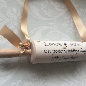 May include: A white wooden rolling pin with a gold ribbon and a peach-coloured flower. The rolling pin is personalised with the text "Louise & Jason On your wedding day 11th June 2016".