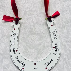 May include: A white horseshoe-shaped ornament featuring red ribbon ties with small red roses at the top. The surface is decorated with dark red dots and black text that reads Mummy and Daddy, 22nd August 2026, with dashed lines along the edges.
