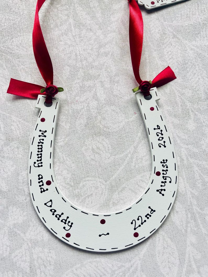 May include: A white horseshoe-shaped ornament featuring red ribbon ties with small red roses at the top. The surface is decorated with dark red dots and black text that reads Mummy and Daddy, 22nd August 2026, with dashed lines along the edges.