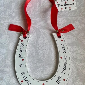 May include: A white horseshoe-shaped ornament featuring red polka dots and black text reading Michael & Roisin and 13th December 2025. It is suspended by a red satin ribbon with small red fabric roses and includes a small white tag reading Love The Horners x.