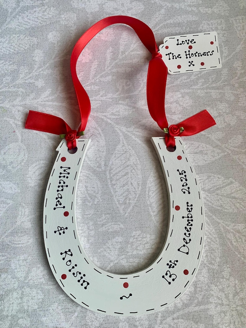 May include: A white horseshoe-shaped ornament featuring red polka dots and black text reading Michael & Roisin and 13th December 2025. It is suspended by a red satin ribbon with small red fabric roses and includes a small white tag reading Love The Horners x.