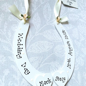 May include: White wooden horseshoe with the words "Wedding Day" and "15th August 2024" written in black script. Two heart-shaped cutouts read "Mark" and "Stacy". A tag reads "Love from Sue x". The horseshoe is decorated with cream-colored ribbon and small floral accents.