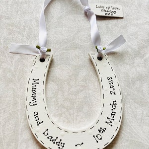 May include: A white wooden horseshoe ornament featuring black hand-lettered text that reads Mummy and Daddy and 10th March 2023. It is suspended by a white satin ribbon with small green fabric roses and includes a small tag reading Lots of love. Stanley xxx.