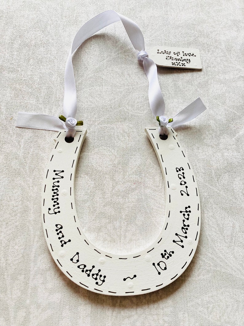 May include: A white wooden horseshoe ornament featuring black hand-lettered text that reads Mummy and Daddy and 10th March 2023. It is suspended by a white satin ribbon with small green fabric roses and includes a small tag reading Lots of love. Stanley xxx.