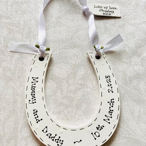 May include: A white wooden horseshoe ornament featuring black hand-lettered text reading Mummy and Daddy and 10th March 2023. It is suspended by a white ribbon with small green fabric roses and includes a small tag labeled Lots of love, Stanley xxx.