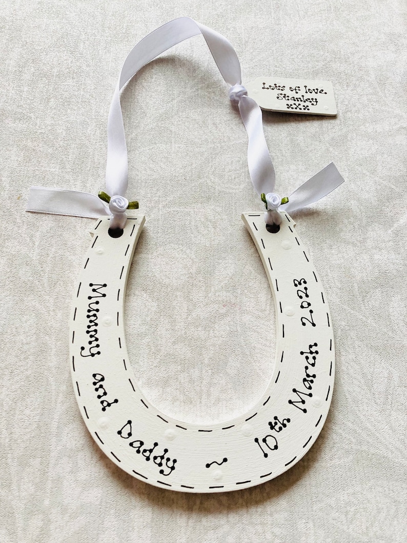 May include: A white wooden horseshoe ornament featuring black hand-lettered text reading Mummy and Daddy and 10th March 2023. It is suspended by a white ribbon with small green fabric roses and includes a small tag labeled Lots of love, Stanley xxx.