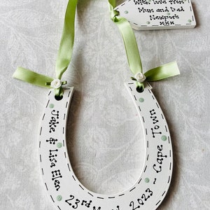 May include: A white wooden horseshoe decoration featuring light green ribbon, small white fabric roses, and black text reading "Jake & Lisa Elen 23rd March 2023 - Cape Town". An attached white wooden tag reads "With love from Mum and Dad Newport xxx".