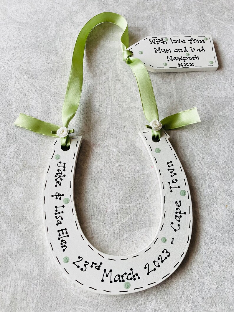 May include: A white wooden horseshoe decoration featuring light green ribbon, small white fabric roses, and black text reading "Jake & Lisa Elen 23rd March 2023 - Cape Town". An attached white wooden tag reads "With love from Mum and Dad Newport xxx".