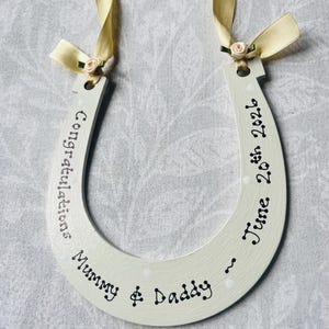 May include: A cream-colored wooden horseshoe ornament featuring black hand-lettered text that reads Congratulations Mummy & Daddy ~ June 20th 2026. The horseshoe is decorated with two pale yellow satin ribbons and small fabric flowers at the top.