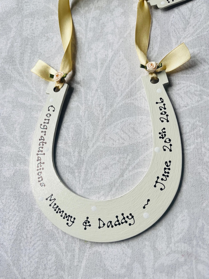 May include: A cream-colored wooden horseshoe ornament featuring black hand-lettered text that reads Congratulations Mummy & Daddy ~ June 20th 2026. The horseshoe is decorated with two pale yellow satin ribbons and small fabric flowers at the top.