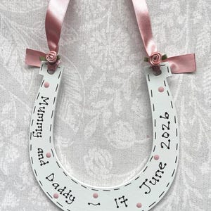 May include: A white horseshoe-shaped wooden ornament featuring pink ribbon loops and small fabric roses at the top. The surface is decorated with black hand-lettered text reading Mummy and Daddy 17 June 2026, accented with small pink painted dots and dashed lines.