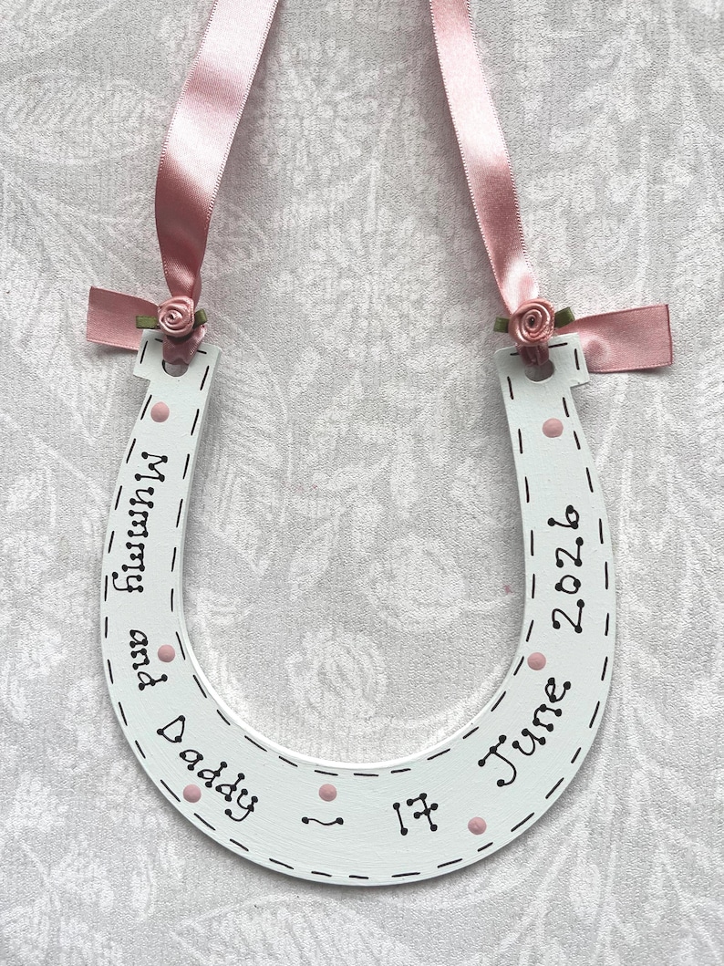May include: A white horseshoe-shaped wooden ornament featuring pink ribbon loops and small fabric roses at the top. The surface is decorated with black hand-lettered text reading Mummy and Daddy 17 June 2026, accented with small pink painted dots and dashed lines.