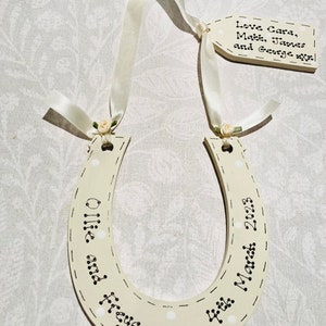 May include: A cream-colored wooden horseshoe ornament featuring white polka dots and black text reading Ollie and Freya 4th March 2023. It hangs from a cream satin ribbon with small fabric roses, attached to a rectangular tag reading Love Cara, Matt, James and George.