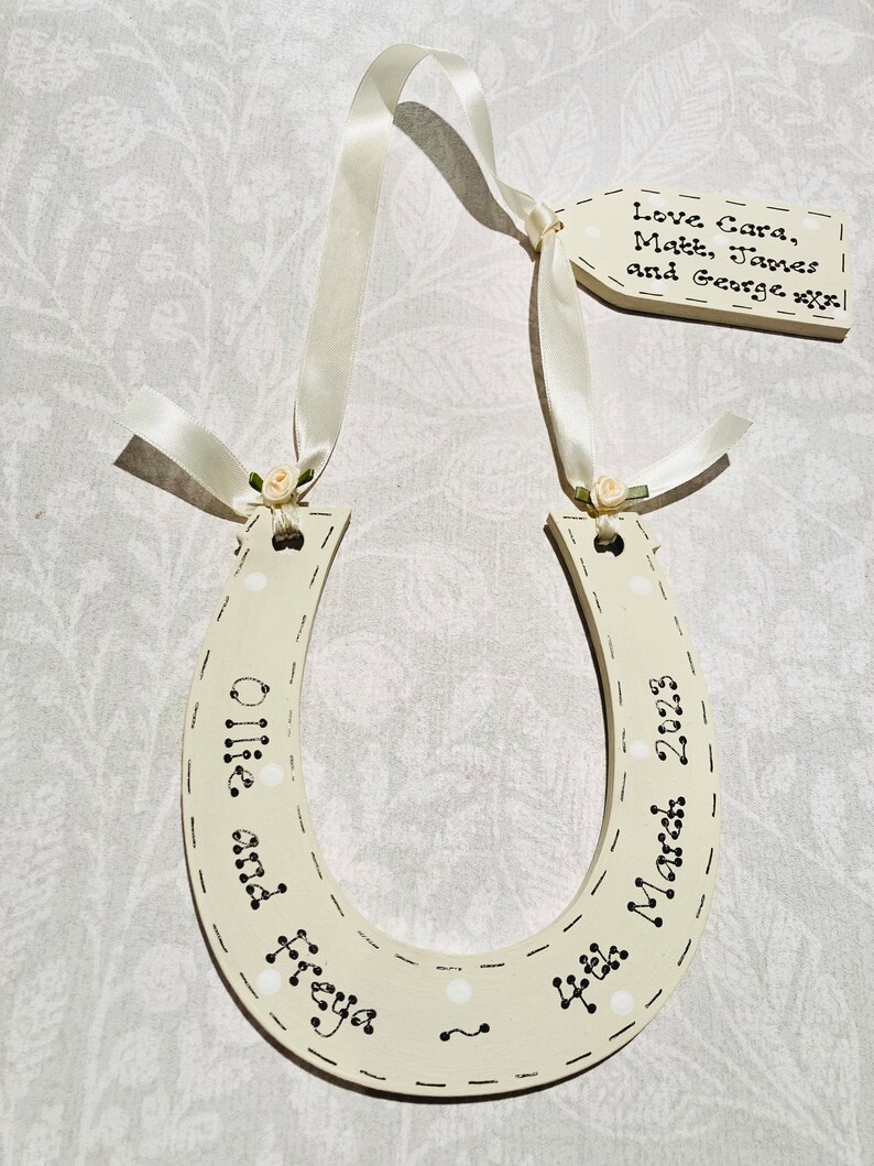 May include: A cream-colored wooden horseshoe ornament featuring white polka dots and black text reading Ollie and Freya 4th March 2023. It hangs from a cream satin ribbon with small fabric roses, attached to a rectangular tag reading Love Cara, Matt, James and George.