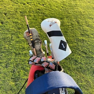 May include: A blue golf bag with a white headcover with the text "Tobacco Road" and a skull and crossbones logo. The headcover has a white number "1" on it. The bag has a red towel and a golf club with a red, white, and blue patterned headcover.