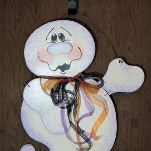 May include: A white ghost with a purple and white painted design, wearing a black and white checkered ribbon and orange, purple, and white ribbons. The ghost has a painted face with green eyes, a pink nose, and a pink blush.