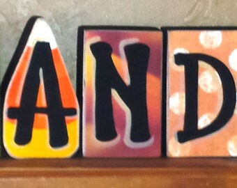 May include: Wooden block letters spelling out the word "CANDY". Each letter is painted black and has a different patterned background. The "C" has a striped background, the "A" has a candy corn pattern, the "N" has a red and orange pattern, the "D" has a white polka dot pattern, and the "Y" has a yellow and orange plaid pattern.