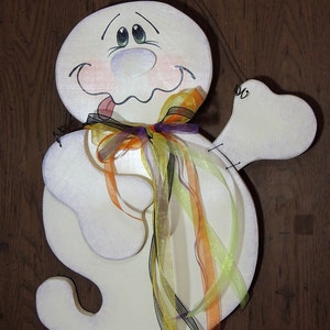 May include: A white ghost with a painted face and a purple, orange, and yellow ribbon bow. The ghost has a tongue sticking out and is holding a white arm out to the side.