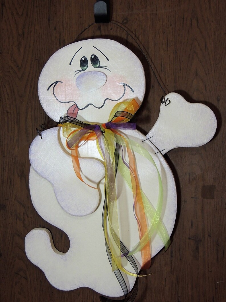 May include: A white ghost with a painted face and a purple, orange, and yellow ribbon bow. The ghost has a tongue sticking out and is holding a white arm out to the side.