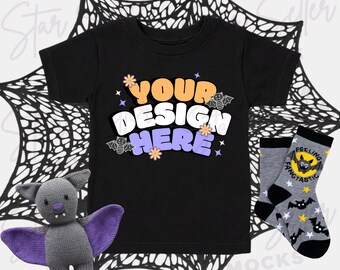 Könnte beinhalten: Ein schwarzes T-Shirt mit dem Text YOUR DESIGN HERE in orangefarbenen, weißen und violetten Buchstaben, verziert mit kleinen Blumen, Sternen und Fledermaus-Illustrationen. Eine graue Fledermaus-Plüschfigur mit violetten Flügeln und graue Socken.