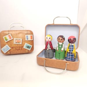 May include: Two miniature suitcases, one with stamps and the word "TRIP", the other open with three wooden peg dolls inside. The dolls are dressed in colorful outfits, suggesting a travel theme. The suitcases are brown with silver handles.