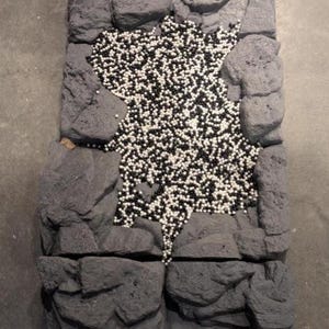 Op de afbeelding: Een rechthoekige, grijze, getextureerde rotswand met een centrale opening gevuld met zwarte en witte bolvormige kralen. De muur bestaat uit vier secties, elk met een ruw, steenachtig uiterlijk.