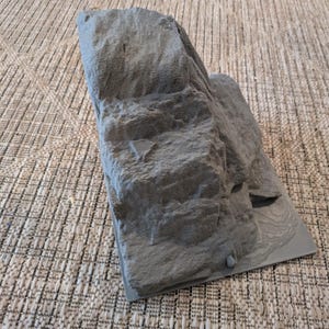May include: A gray, textured miniature rock formation, likely for a model or diorama. The rock has a rough, uneven surface with visible details. It sits on a flat, gray base.