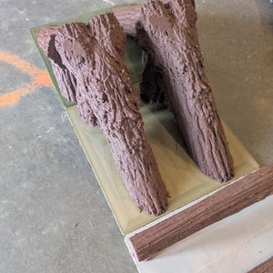 Peut inclure: Deux supports en forme de bûche, imprimés en 3D, de couleur brune et texturés, fixés sur une base plate rectangulaire de couleur claire. Une pièce séparée, longue, fine, brune et texturée, repose sur une surface blanche au premier plan.